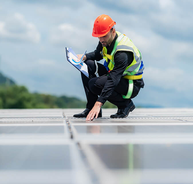 Technician kneeling on a rooftop solar array while recording inspection notes on a clipboard